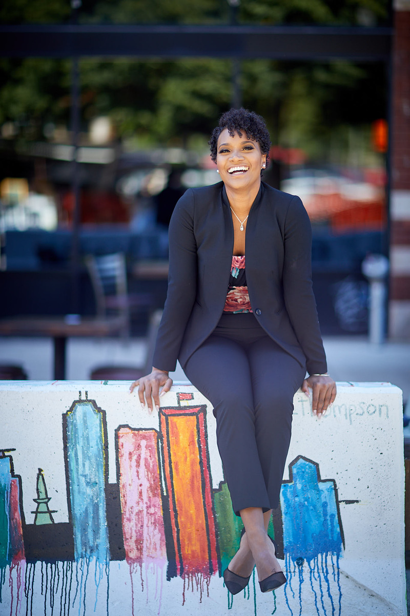 Portrait of a woman in a black suit laughing while seated on a low wall painted with a colourful city skyline mural